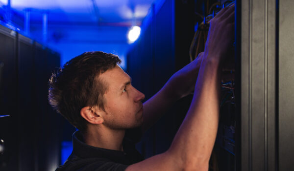 Close-up of a young, light-skinned man in a dark shirt, arms raised, focused on manipulating cables and equipment inside a brightly lit server rack in a dark data center.